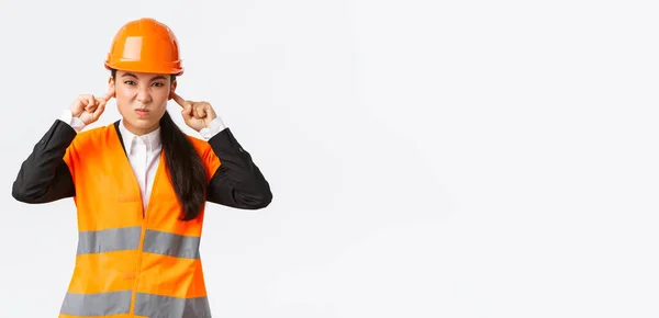 Inquiète et mécontente asiatique chef mécanicienne fermer les oreilles et grimaçant de bruit terriblement fort dans la zone de construction, portant un casque de sécurité, se plaignant sur le bruit dérangeant, fond blanc — Photo