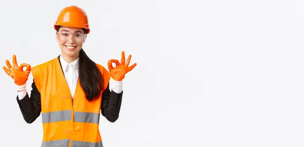 Zelfverzekerde aziatische vrouwelijke bouwkundig ingenieur, enterprise manager tonen oke gebaar na het aantrekken van veiligheidshelm, bril en handschoenen voor het invoeren van gevaarlijke gebied, witte achtergrond — Stockfoto