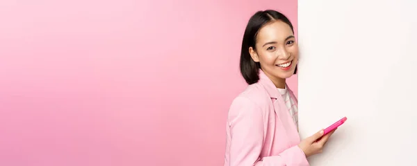 Imagem de empresária coreana de terno, de pé perto da parede info, propaganda a bordo, segurando smartphone e sorrindo, posando sobre fundo rosa — Fotografia de Stock