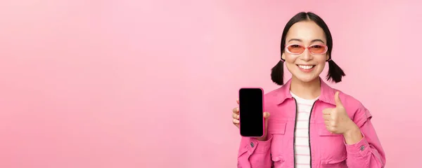 Счастливая стильная девушка запускает приложение на мобильном телефоне. Улыбающаяся азиатская модель со смартфоном и большим пальцем, стоящая на розовом фоне — стоковое фото