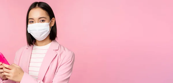 Asian businesswoman in medical face mask using mobile phone. Japenese saleswoman, corporate lady in suit, holding smartphone, standing over pink background — Stock Photo, Image