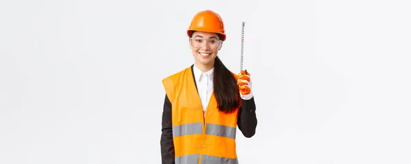 Впевнений усміхнений, професійний азіатський інженер-жінка, технік будівництва в шоломі безпеки та рефлекторній формі, стоїть зі стрічковою мірою, вимірюючи планування на будівельній ділянці — стокове фото