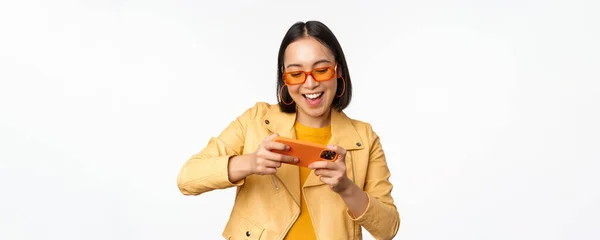 Stylowa koreańska dziewczyna w okularach przeciwsłonecznych, grająca w mobilną grę wideo, śmiejąca się i uśmiechnięta podczas korzystania ze smartfona, stojąca na białym tle — Zdjęcie stockowe