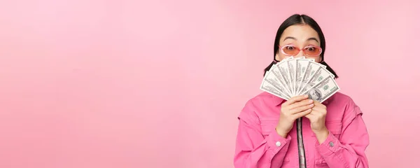 Belle coréenne en lunettes de soleil, montrant des dollars, argent comptant, sourire heureux, concept de prêts rapides, microcrédit et paiement, debout sur fond rose — Photo