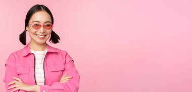 Pembe elbiseli ve güneş gözlüklü şık Asyalı kız, kameraya gülümsüyor ve mutlu görünüyor, stüdyo arka planında duruyor.