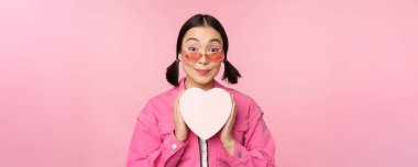 Güzel Asyalı kız mutlu gülümsüyor, kalp hediye kutusunu gösteriyor ve kameraya heyecanlı bakıyor, pembe romantik arka planda duruyor.