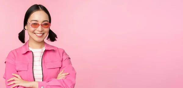 Стильна азіатська дівчина в рожевому одязі і сонцезахисних окулярах, посміхаючись і дивлячись щасливо на камеру, стоїть над студійним фоном — стокове фото