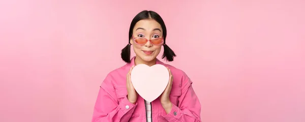 Hermosa chica asiática sonriendo feliz, mostrando caja de regalo de corazón y mirando emocionada a la cámara, de pie sobre fondo romántico rosa —  Fotos de Stock