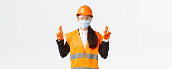 Covid-19肠胃安全规程，构建和预防病毒概念。有希望的亚洲女工程师头戴头盔，戴口罩，祈祷，闭上眼睛，交叉手指，好运，白色背景 — 图库照片