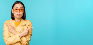 Güneş gözlüklü kararsız Asyalı kadın, iki varyant arasında seçim yapıyor, iki yana bakıyor, şaşkın görünüyor, mavi arka planda duruyor.