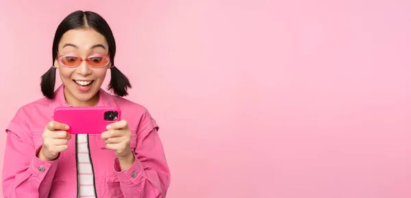 Portret van gelukkig aziatisch meisje spelen op smartphone, kijken naar video 's op mobiele telefoon app, staande over roze achtergrond — Stockfoto