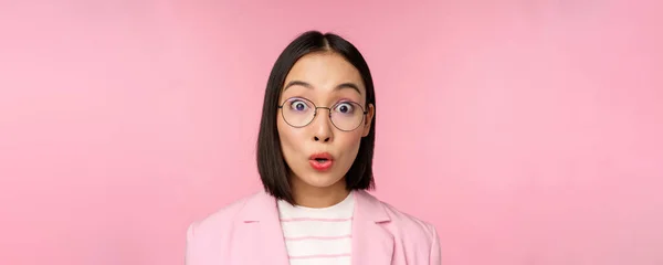 Primer plano retrato de asiático mujer de negocios en gafas mirando sorprendido en cámara, sorprendido reacción, de pie en traje sobre rosa fondo —  Fotos de Stock