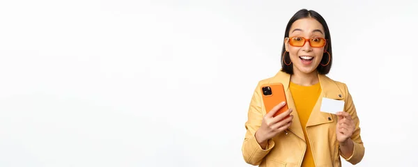 Compras en línea. Elegante modelo femenina asiática en gafas de sol, celebración de la tarjeta de crédito y el teléfono móvil, sonriendo feliz, de pie sobre fondo blanco — Foto de Stock