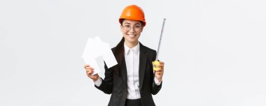 Gülümseyen Asyalı kadın mühendis, miğfer ve iş elbisesi içinde ev maketi ve mezurayı gösteren mimar, ev yenilemesine, inşaat çalışmalarına, beyaz arka plana başlamaya hazır.