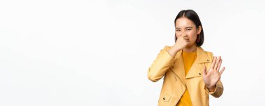 Asyalı kız görüntüsü burnunu kapatıyor ve iğrenç bir kokuyla smth 'i reddediyor, burnunu kapatıyor, beyaz arka planda duruyor.