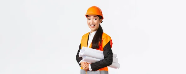Incrível e animado sorrindo asiático arquiteto feminino, engenheiro-chefe em capacete de segurança e revestimento reflexivo olhando divertido e feliz, sorrindo impressionado, segurando plantas de área de construção — Fotografia de Stock