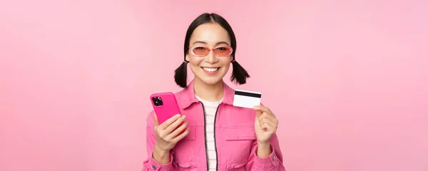 Zakupy online. Uśmiechnięta azjatycka dziewczyna shopper, trzymając smartfon i kartę kredytową, płacąc w aplikacji mobilnej, stojąc na różowym tle — Zdjęcie stockowe