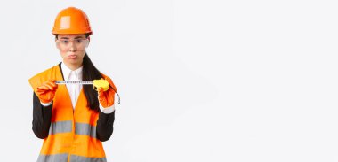 Hüzünlü Asyalı kadın inşaat mühendisi, mimar mezurada küçük boy gösteriyor, ölçümlerden rahatsız, güvenlik kaskından hoşnutsuz, beyaz arka plan.