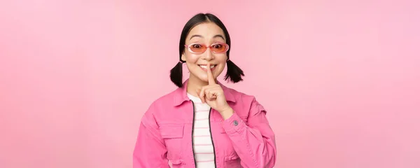 Chica coreana moderna en traje de primavera con estilo, gafas de sol, mostrando el silencio, signo de silencio, presionar el dedo a los labios, gesto tabú, de pie sobre el fondo rosa — Foto de Stock