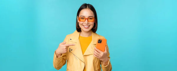 Junge stylische asiatische Model in trendigen Sonnenbrille, Frühling Outfit, zeigt Handy, zeigt auf Smartphone und lächelt, steht über blauem Hintergrund — Stockfoto