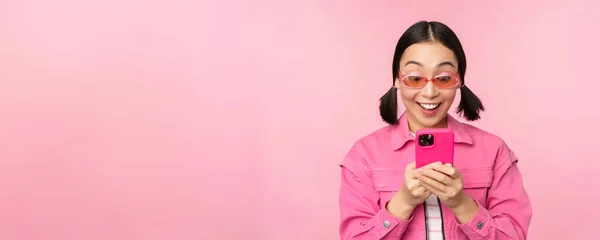 Ritratto di ragazza asiatica in occhiali da sole con smartphone. Donna guardando il telefono cellulare, navigando in app, in piedi su sfondo rosa — Foto Stock