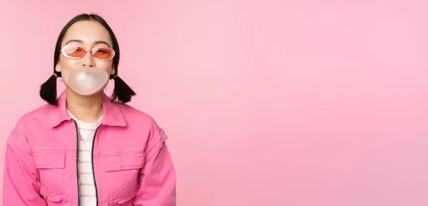 Стильная азиатская девушка, надувающая пузырь жвачки, жующая жвачку, в солнечных очках, позирующая на розовом фоне — стоковое фото