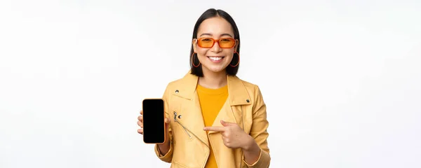 Ritratto di elegante ragazza coreana in occhiali da sole, sorridente, puntare il dito contro lo schermo dello smartphone, mostrando l'applicazione del telefono cellulare, in piedi su sfondo bianco — Foto Stock