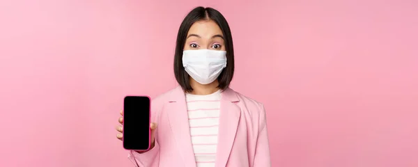 Retrato de vendedora coreana sorridente em máscara facial médica, terno de negócios, mostrando tela do smartphone, de pé sobre fundo rosa — Fotografia de Stock