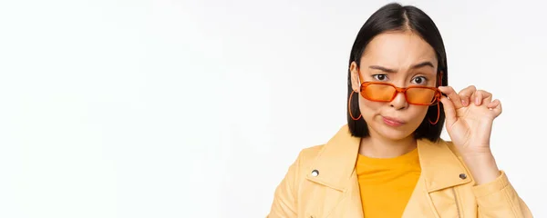 Imagen de mujer joven asiática con estilo, mirando desde debajo de las gafas de sol con cara escéptica, incredulidad, duda expresa, de pie sobre fondo blanco — Foto de Stock