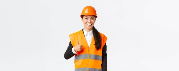 Magabiztos elégedett ázsiai női építőipari vezető a biztonsági sisak, fényvisszaverő kabát, mutatja hüvelykujj-up jóváhagyása, támogatás és hasonló munka az alkalmazottak, álló fehér háttér — Stock Fotó