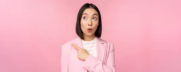 Portrait of saleswoman, korea business woman pointing and looking left with surprised, incregued face expression, posing in suit over pink background — Stok Foto