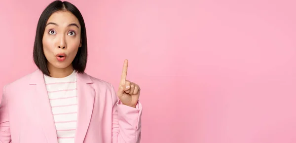 Entusiasta trabajadora corporativa, mujer de negocios asiática señalando con los dedos hacia arriba y sonriendo, mostrando publicidad, logotipo, de pie sobre fondo rosa —  Fotos de Stock