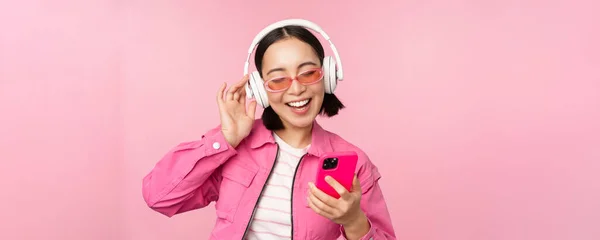 Стильна азіатська дівчина танцює зі смартфоном, слухаючи музику в навушниках на мобільному телефоні, посміхаючись і сміючись, позує на рожевому фоні — стокове фото