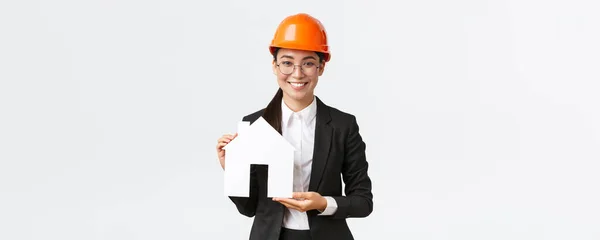 Sonriente mujer atractiva vendedora asiática de la empresa de construcción que vende casas, usa casco y traje de negocios, mostrando maket de casa acogedora, de pie fondo blanco — Foto de Stock