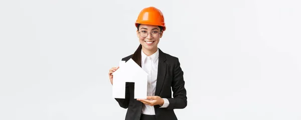 Souriant belle femme asiatique vendeuse d'entreprise de construction vendant des maisons, porter un casque et costume d'affaires, montrant maket de maison confortable, debout fond blanc — Photo