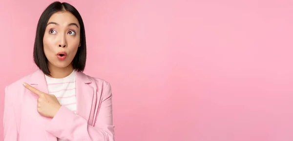 Retrato de vendedora, empresaria coreana señalando y mirando a la izquierda con expresión de cara sorprendida e intrigada, posando en traje sobre fondo rosa —  Fotos de Stock