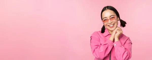 戴着太阳镜的时髦亚洲女人的近照，面带微笑，面带微笑，笑容可亲，站在粉色背景上 — 图库照片