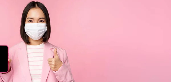 Portrait of asian corporate woman in medical face mask covid-19, showing smartphone screen and thumb up sign, pink background — Stock Photo, Image