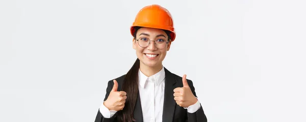 Close-up of professional pleased asian female chief engineer, architect in business suit and safety helmet showing thumbs-up in approval, give permission, recommend construction company — Stock Photo, Image