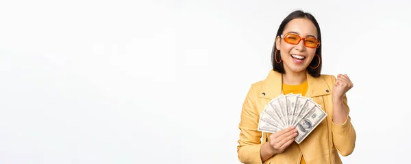 Stylish smiling asian girl holding money cash, showing dollars and celebrating, standing over white background — Stock Photo, Image