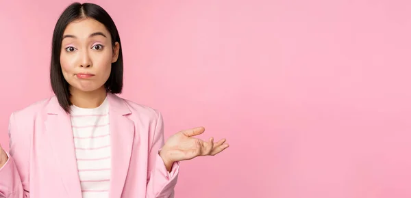 Πορτρέτο της μπερδεμένος ασιατική επιχειρηματίας shrugging ώμους, αναζητούν ανίδεοι και προβληματισμένος, δεν ξέρω, δεν μπορώ να πω, στέκεται πάνω από ροζ φόντο στο κοστούμι γραφείου — Φωτογραφία Αρχείου