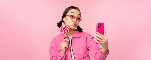 Portret van stijlvol, gelukkig aziatisch meisje dat selfie maakt met snoep, lolipop snoep en glimlach, foto 's maakt met mobiele app, over een roze achtergrond staat — Stockfoto