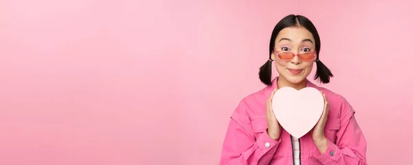 Linda menina asiática sorrindo feliz, mostrando caixa de presente do coração e olhando animado para a câmera, de pé sobre o fundo romântico rosa — Fotografia de Stock