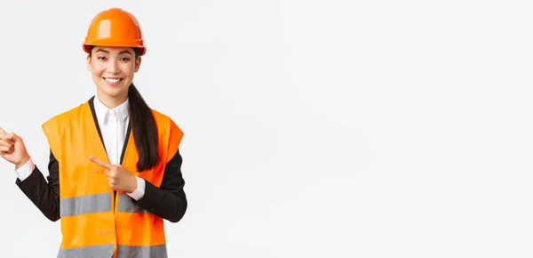 Construcción, construcción y concepto industrial. Sonriente asiática arquitecta en casco de seguridad, ropa reflectante apuntando dedo esquina superior izquierda, mostrando proyecto en el lugar de trabajo, fondo blanco — Foto de Stock