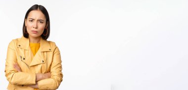 Asyalı bir kızın kızgın ve incinmiş görünümü, üzgün, gücenmiş yüz ifadesi, beyaz arka planda dikilmesi.