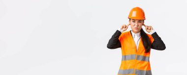 Rahatsız ve hoşnutsuz Asyalı kadın mühendis kulaklarını kapattı ve inşaat alanındaki korkunç gürültüden dolayı yüzünü buruşturdu, güvenlik kaskı taktı, rahatsız edici sesten şikayet etti, beyaz arka plan