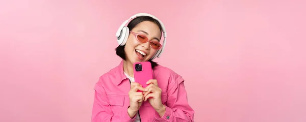 Stylowa azjatycka dziewczyna w słuchawkach, słuchanie muzyki i robienie zdjęć przez telefon komórkowy, za pomocą smartfona, stojąc nad różowym tle — Zdjęcie stockowe
