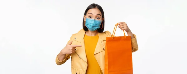 Covid-19 és üzletkoncepció. Fiatal ázsiai stílusos nő, orvosi maszkot visel, kezében bevásárló táska, megy a plázákban világjárvány idején, fehér háttér — Stock Fotó