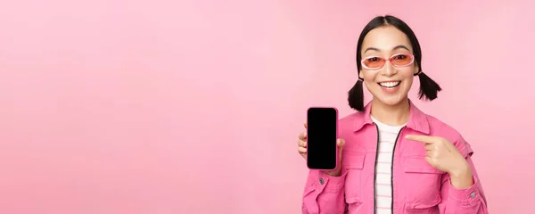 Enthousiaste Aziatische vrouw in stijlvolle kleding, zonnebril, wijzende vinger naar mobiele telefoon scherm, tonen smartphone applicatie, staande over roze achtergrond — Stockfoto