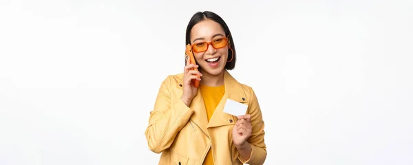 Elegante modelo femenino asiático, hablando en el teléfono inteligente y mostrando la tarjeta de crédito, de pie sobre fondo blanco — Foto de Stock